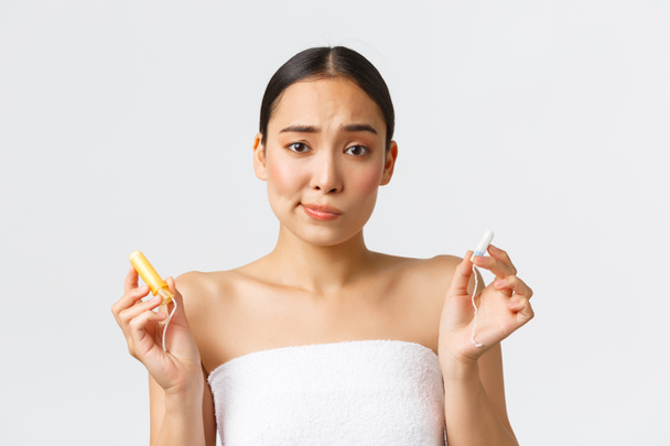 Beauty, personal and intimate care menstrual hygiene concept. Cute indecisive asian girl on her perion standing in towel and holding tampons with and without applicator, dont know what choose - Photo, Image