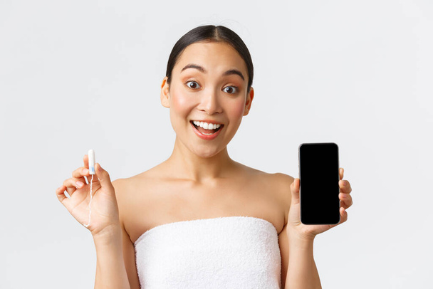 Schönheit, persönliche und intime Pflege, mobiles Anwendungskonzept. Fröhlich lächelnde Asiatin zeigt ihren Menstruationszyklus, Zeitraffer auf Smartphone-Bildschirm, hält Tampon in der Hand - Foto, Bild