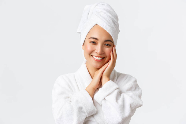 Cuidados pessoais, mulheres beleza, banho e chuveiro conceito. Close-up de bela mulher asiática feliz em toalha e roupão tocando rosto suavemente, sorrindo dentes brancos, promo de cuidados com a pele e produtos de higiene
 - Foto, Imagem