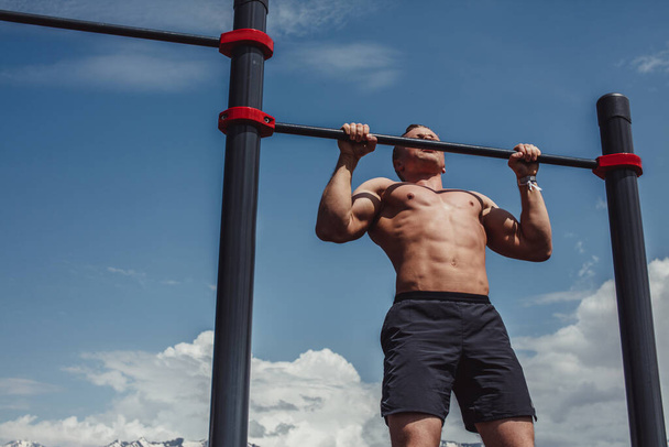 Sports man doing pull-up exercise on a horizontal bar against a blue sky. - 写真・画像