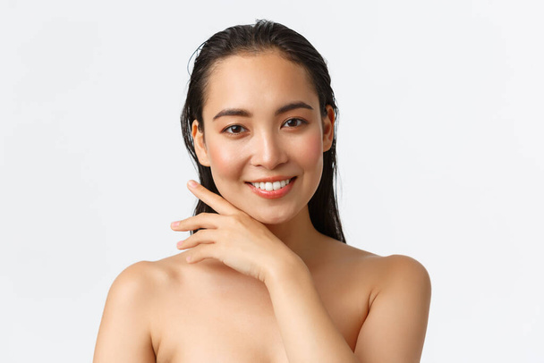 Skincare, bodycare, beauty and bath concept. Close-up of sensual attractive asian woman standing naked in shower, smiling and gently touching face, promo of facial products, white background - Photo, Image