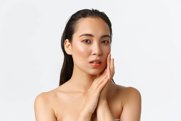 Skincare, bodycare, beauty and bath concept. Sensual beautiful asian woman standing naked, touching face gently and looking dreamy camera, apply facial cream, nourishing mask - Photo, Image