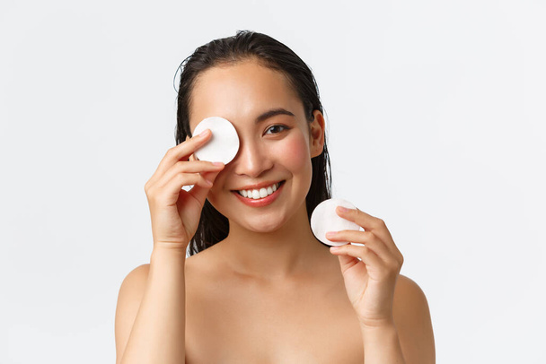 Skincare, women beauty, hygiene and personal care concept. Close-up of attractive naked asian woman in bathroom, wipe-off makeup with cotton pads and smiling camera, white background - Фото, изображение