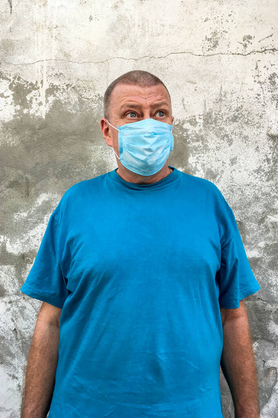 Kurzhaariger kaukasischer Ethnizität reifer Mann in blauem T-Shirt mit medizinischer Maske auf dem Gesicht, um COVID-19 zu schützen, mit Blick zur Seite vor dem Hintergrund einer grau verputzten Wand. Vertikaler Schuss. Nahaufnahme. - Foto, Bild