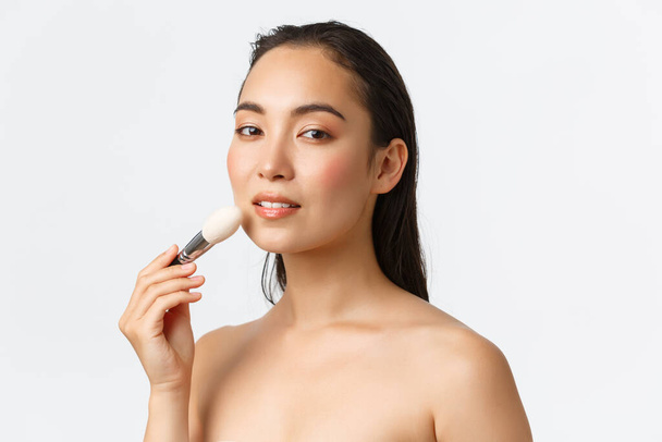 Skincare, women beauty, hygiene and personal care concept. Sensual beautiful asian woman standing naked with makeup brush, looking camera seductive, white background - Foto, Bild