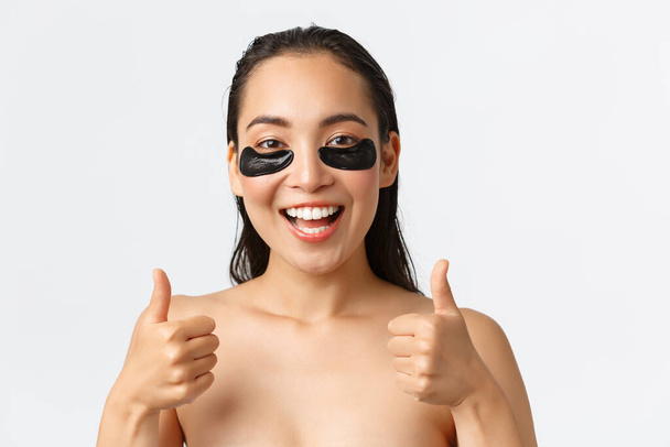 Cuidados com a pele, beleza feminina, higiene e conceito de cuidados pessoais. Close-up de satisfeito atraente asiático mulher tratamento de sacos de olho com sob os olhos remendos, mostrando polegares-para cima, de pé nu
 - Foto, Imagem