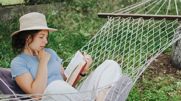 Thoughtful young woman in summer straw hat reading interesting book in a hammock in green garden during weekend.Curly brunette girl student absorbed in reading best seller sitting on rope hummock. - Photo, Image