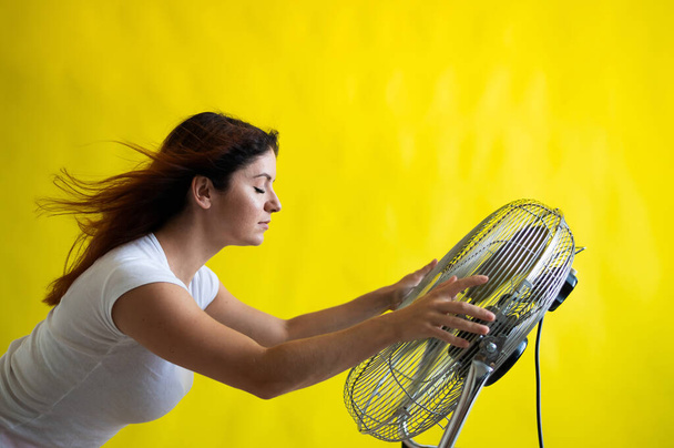 A beautiful red-haired woman is cooled off standing over a large electric fan on a yellow background. Girl with hair developing in the wind. Device for cooling the air. - Foto, afbeelding