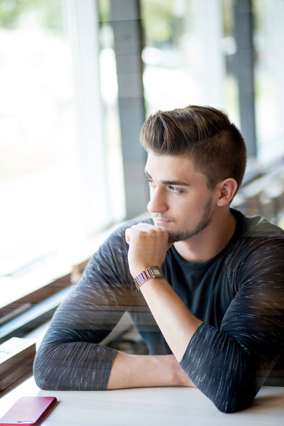 Young handsome man sitting near window at street cafe with pensive expression. - 写真・画像