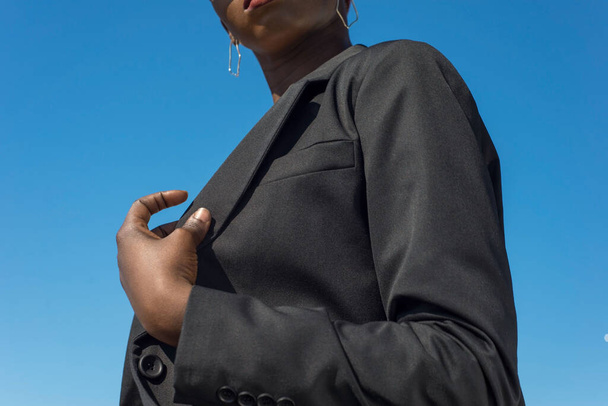 Молодая африканская модная деловая женщина в черном деловом пиджаке и брюках, позирующих с деталями костюма. На фоне голубого неба. Стиль деловой моды в черном костюме, на черной девушке. Африканский стиль бизнеса - Фото, изображение