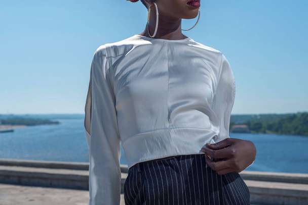 молодая африканская черная модель, изображающая детали белой блузки. Модный выстрел в голубое небо. Рекламное фото белой женской блузки на черной девушке-модели. женский манекен в белой блузке на белом фоне - Фото, изображение