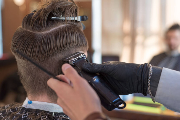 美容室が髭を生やした若い男を髪の毛のトリマーで切り取って頭に毛をつけてる。理髪店でのメンズヘアカットの巨匠の仕事 - 写真・画像