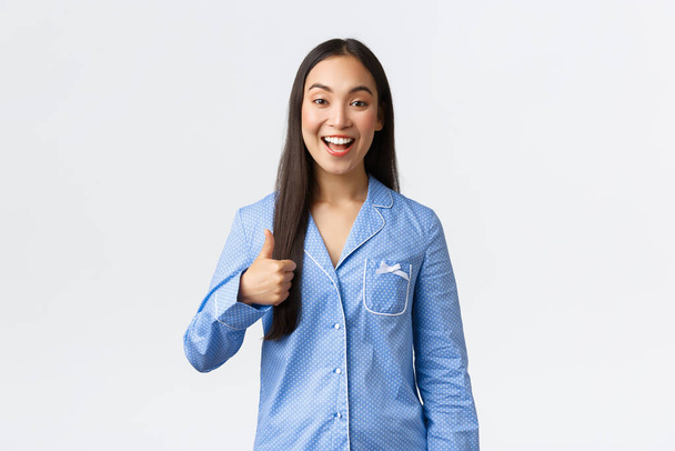 Optimistisch glücklich asiatische hübsche Mädchen im blauen Pyjama lächelt zufrieden und zeigt Daumen nach oben in Zustimmung oder wie, empfehlen Produkt, gute Qualität, zeigt gut gemacht oder gute Arbeit, weißer Hintergrund - Foto, Bild