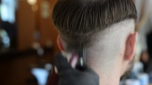 een kapper knipt een bebaarde jongeman met een haarknipper, kamt het haar op zijn hoofd. Werk van de meester in mannen kapsel in een kapperszaak - Video