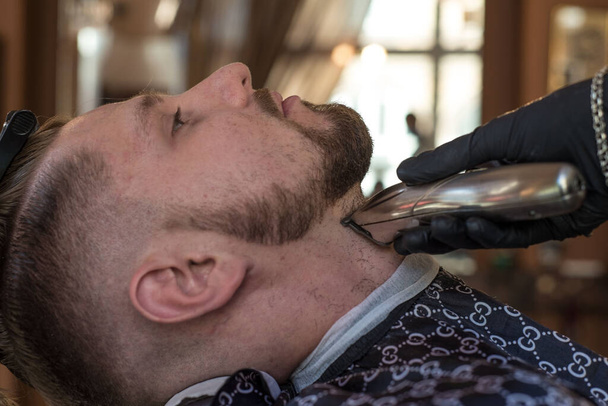 перукар розрізає бородатого молодого хлопця тримером для волосся, розчісуючи волосся на голові. Робота майстра в чоловічій стрижці в перукарні
. - Фото, зображення