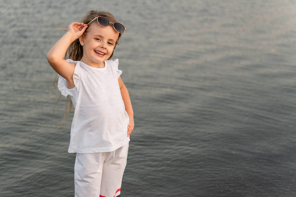 Photo of a Beautiful little girl on the background of a pond with copy space. A girl in a white suit against the background of a calm sea. - Foto, Imagen