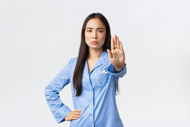Mavi pijamalı ciddi görünümlü Asyalı kız dur demek için elini uzatıyor, eylemi yasaklıyor, uyarı yapıyor ve kötü davranışı onaylamıyor, beyaz arka planda bir şeyi yasaklıyor. - Fotoğraf, Görsel