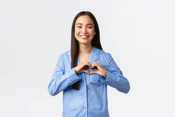 schattig teder aziatisch meisje in blauwe pyjama tonen hart gebaar en glimlachen, uiten liefde en zorg, belijden sympathie, staan in pyjama houden van geweldig logeerpartijtje, witte achtergrond - Foto, afbeelding