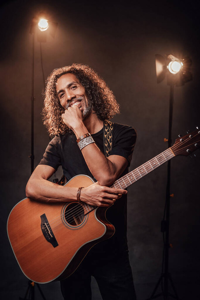 Hispánec středního věku v černém tričku drží kytaru na scéně, usmívá se a dívá se do kamery - Fotografie, Obrázek