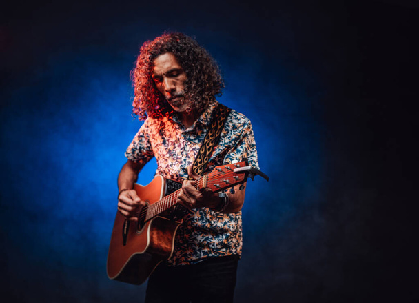 ハワイのシャツに身を包んだ才能あるヒスパニックミュージシャンが暗闇でギターを弾く - 写真・画像