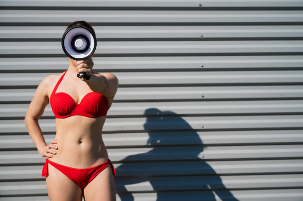 Portrait of a woman in a red bikini holds a megaphone on the background of the face. A girl in a swimsuit makes an announcement on the speaker against the background of a steel sheet. - Valokuva, kuva
