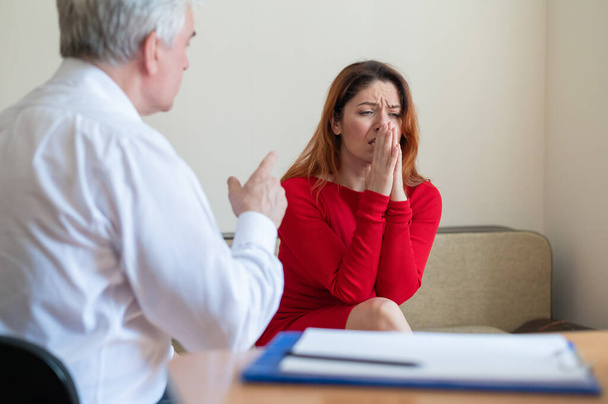 A frustrated crying woman holds her palms over face at a session with a male psychologist. Mature psychotherapist talking to a female patient with depression and neurosis. Mental health treatment. - Фото, изображение