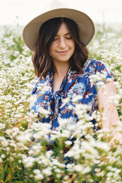 Elegante mujer joven en vestido vintage azul y sombrero sonriendo en el prado de verano en muchas flores silvestres blancas. Tranquilo verano en el campo. Hermosa chica feliz de pie en margaritas blancas
 - Foto, imagen