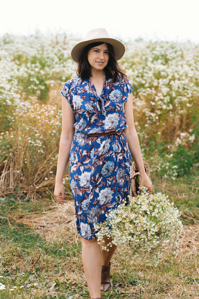 Стильная молодая женщина в синем винтажном платье и шляпе, собирающая белые полевые цветы в соломенной корзине в медоу. Красивая девушка с белым букетиком маргаритки. Спокойное лето за городом - Фото, изображение