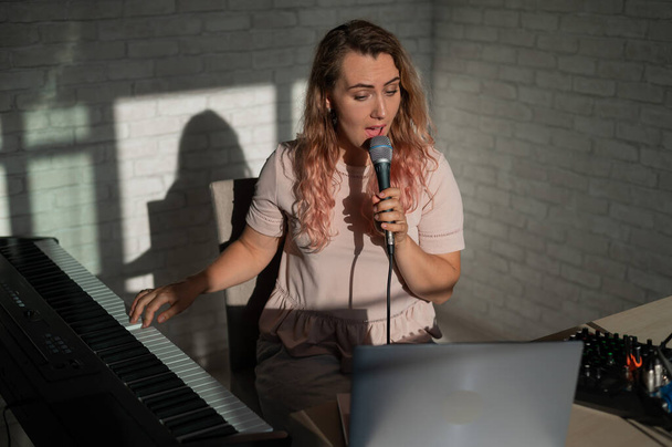 A young European woman sings into a microphone and accompanies on an electronic piano. The girl shoots a video blog. Remote vocal lesson. A female singing teacher conducts online classes. - Fotoğraf, Görsel