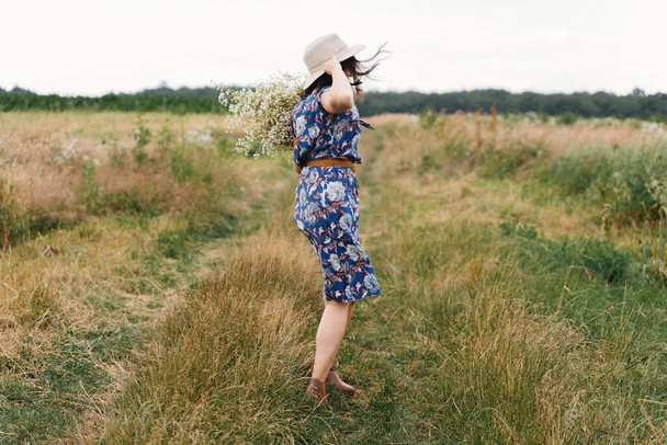 Jovem mulher de vestido vintage azul e chapéu dançando com flores silvestres brancas no prado. Menina bonita se divertindo e rindo no campo ventoso com grande buquê margaridas. Verão autêntico no campo
 - Foto, Imagem