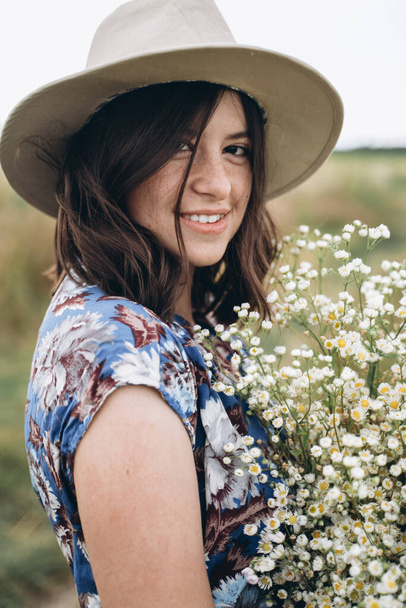 Gyönyörű fiatal nő kék évjárat ruha és kalap kezében fehér vadvirágok a réten. Érzéki portré gyönyörű lány nagy százszorszépek csokor szeles területen. Vidéki nyár - Fotó, kép