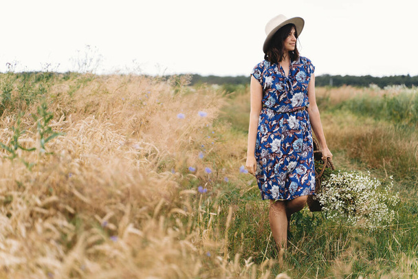 Nyugodt nyár vidéken. Stílusos fiatal nő kék évjárat ruha és kalap séta fehér vadvirágok szalmakosárban sárga búza mező. Gyönyörű lány fehér százszorszépek csokor. - Fotó, kép