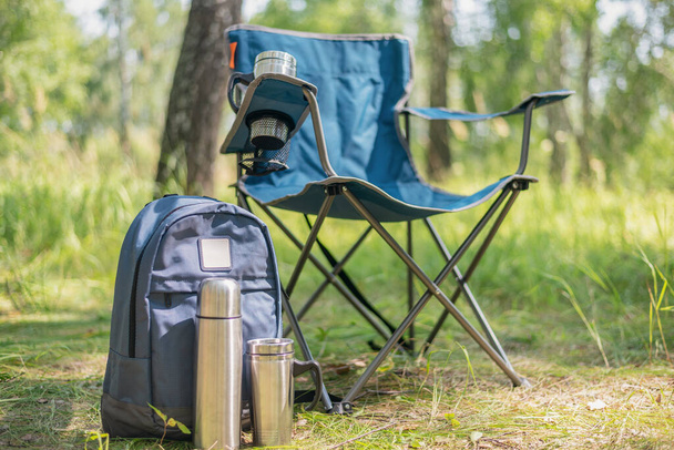 Backpack, vacuum flask, touristic chair and steel cup on the ground on the green forest background. - Foto, Imagem