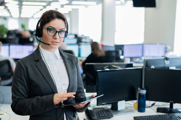 Schöne Kaukasierin mit Headset hält ein digitales Tablet in der Hand, während sie im Großraumbüro steht. Eine freundliche Servicemitarbeiterin surft am Arbeitsplatz auf dem Bildschirm des Geräts. - Foto, Bild