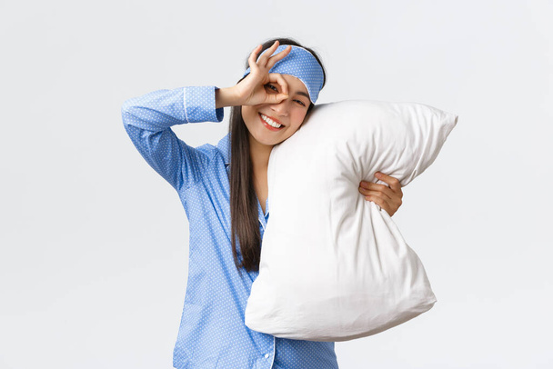 Freudig enthusiastische schöne asiatische Mädchen in blauem Pyjama und Schlafmaske, umarmt Kissen und lächelt optimistisch wie schlafen gehen, bereit zum Schlafen, zeigt okay Geste in Zustimmung, weißer Hintergrund - Foto, Bild