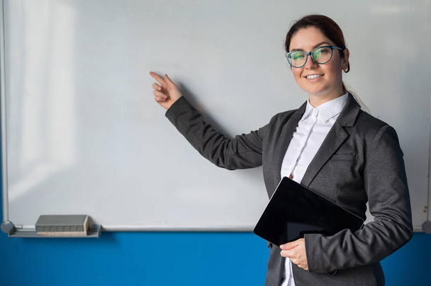 Beautiful woman in a suit with a digital tablet in her hands. A smiling female business coach with glasses stands in a conference room and points a finger at a white board. Workshop at the office. - Zdjęcie, obraz