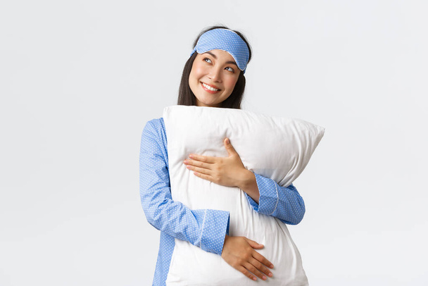 Мрійлива красива азіатська дівчина в блакитній піжамі і спальна маска обіймає подушку і усміхається задоволена, як виглядає верхній лівий кут зображення щось в ліжку, фотографування, використовуючи уяву
 - Фото, зображення