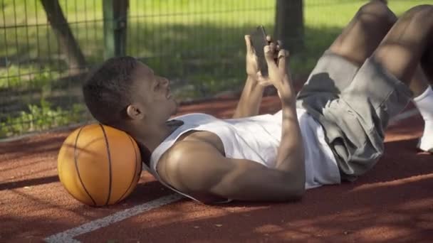 Positive young sportsman taking selfie as lying on ball on basketball court. Side view portrait of cheerful African American player resting outdoors on sunny day. Sport, lifestyle, enjoyment, rest. - Video