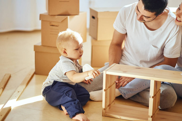 family together at living room of new apartment assembling furniture, pile of moving boxes on background - 写真・画像