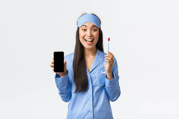 Гігієна, технології та люди вдома концепція. Усміхнена приваблива азіатська дівчина в блакитній піжамі та масці для сну, що показує додаток для навчання дітей, як користуватися зубною щіткою та чистити зуби, тримайте смартфон
 - Фото, зображення