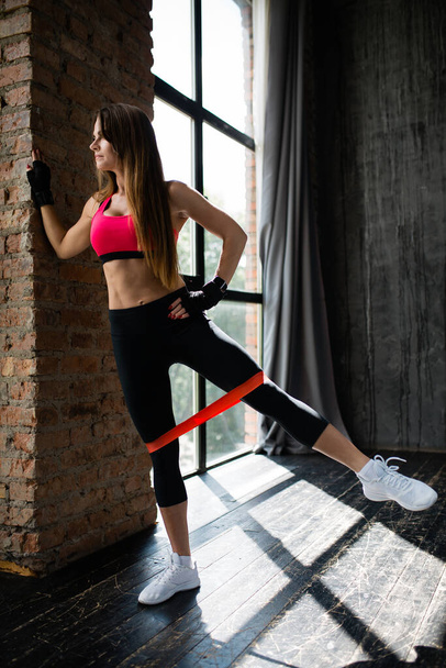 Pilates trainer in a pink top does exercises with an elastic rubber band put on her legs and looks out of the panoramic window in the loft. - Foto, immagini