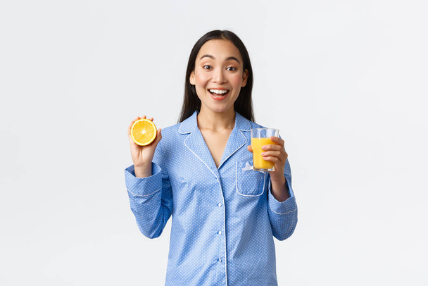 Morgendliches, aktives und gesundes Lebens- und Wohnkonzept. Fröhlich lächelndes asiatisches Mädchen, das ihren Tag mit frisch gemachtem Orangengeschmack beginnt, Glas und halbe Orange in der Hand hält, glücklich und energiegeladen aussieht - Foto, Bild