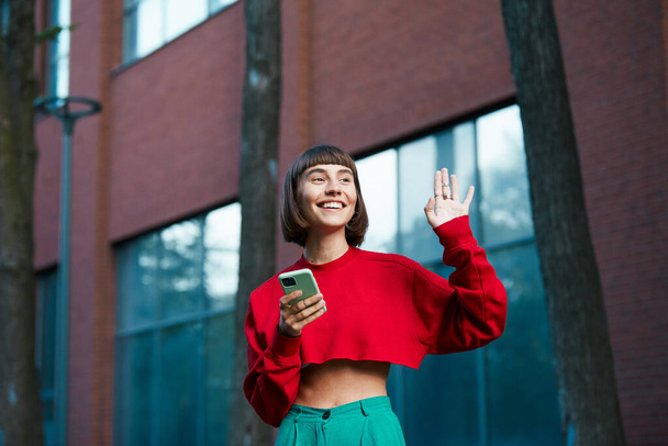 beautiful young girl saying hi on the street and holding phone, cute millenial woman in red stylish sweater having smartphone in arm and showing hi gesture - Фото, изображение