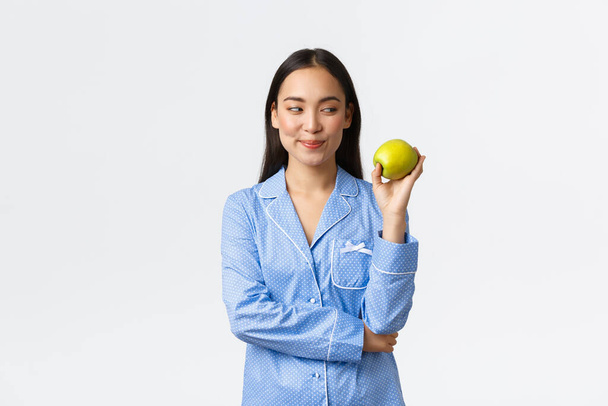 Morgendliches, aktives und gesundes Lebens- und Wohnkonzept. Schlaue schöne asiatische Frau im blauen Pyjama schaut nachdenklich auf grünen Apfel, lächelt verschmitzt, als stünde sie vor weißem Hintergrund - Foto, Bild