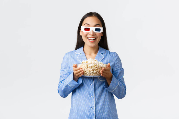 Home leisure, sleepover and slumber party concept. Enthusiastic asian girl in pajamas and 3d glasses, holding bowl of popcorn and smiling amused as watching premier on tv, enjoying movie night - Photo, Image