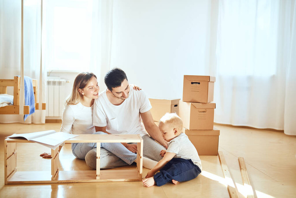 family together at living room of new apartment assembling furniture, pile of moving boxes on background - Фото, изображение