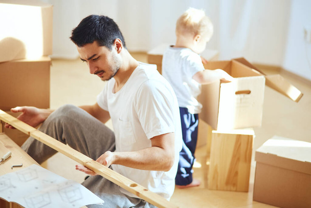 father assembling furniture in living room of new apartment, mother with son and pile of moving boxes on background - Fotografie, Obrázek