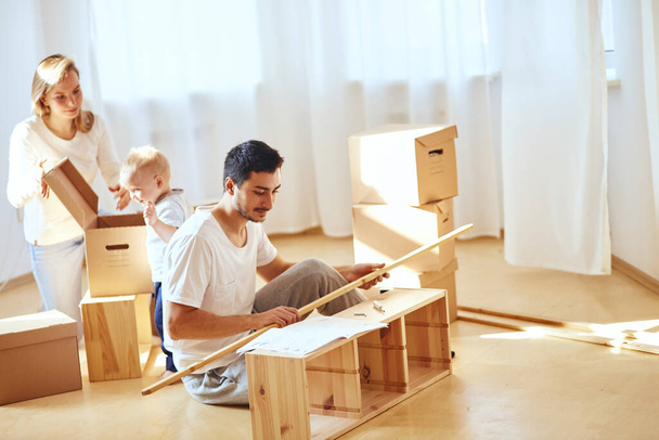 father assembling furniture in living room of new apartment, mother with son and pile of moving boxes on background - 写真・画像