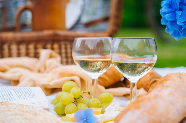 Letni piknik w słoneczny dzień z chlebem, owocami, kwiatami hortensji bukietowej, kieliszkami wina, słomkowym kapeluszem, książką i ukulele. Kosz piknikowy na trawie z jedzeniem i piciem na białym kocu z dzianiny. Skupienie selektywne - Zdjęcie, obraz