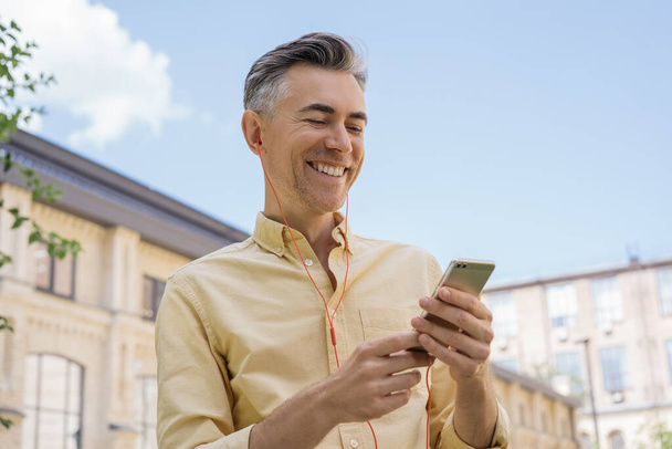 Guapo hombre maduro sosteniendo el teléfono móvil, escuchando música en auriculares rojos, mirando a la pantalla digital. Hombre de negocios sonriente usando el teléfono celular, comprando en línea. Teléfono móvil
 - Foto, imagen